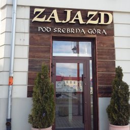 Studio Reklamy RED DOG - Elewacja budynku z szyldem 'Zajazd Pod Srebrną Górą' nad wejściem, ozdobiona drewnianymi panelami i dwoma tujami w glinianych donicach po bokach drzwi.