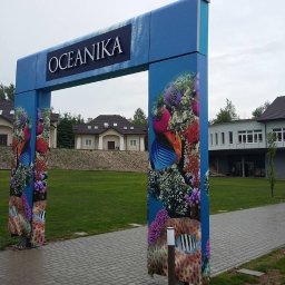 Studio Reklamy RED DOG - Brama wejściowa z niebieską konstrukcją ozdobioną grafiką rafy koralowej i ryb, nad którą umieszczony jest szyld z napisem 'OCEANIKA', prowadząca do budynku w tle.