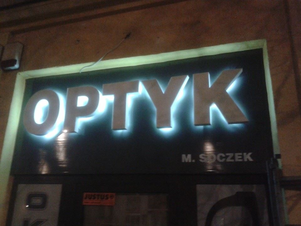 Podświetlany szyld sklepu optycznego 'OPTYK M. SOCZEK' z widocznymi elementami wystawy za szybą.