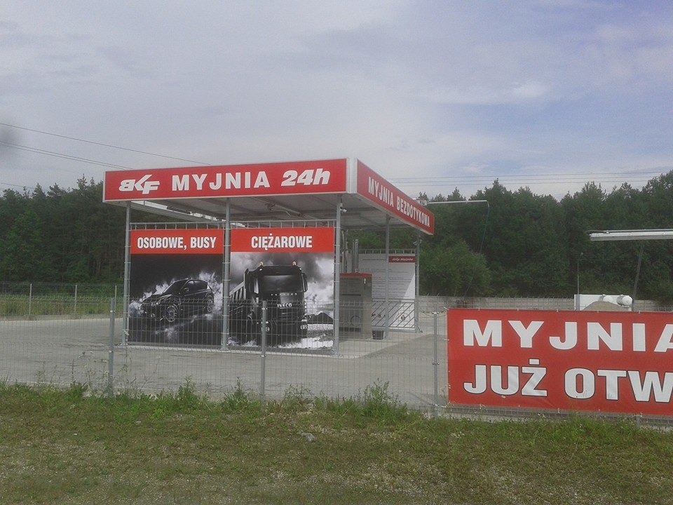 Bezdotykowa myjnia samochodowa 24h z reklamami samochodów osobowych i ciężarowych, widoczna zza metalowego ogrodzenia.