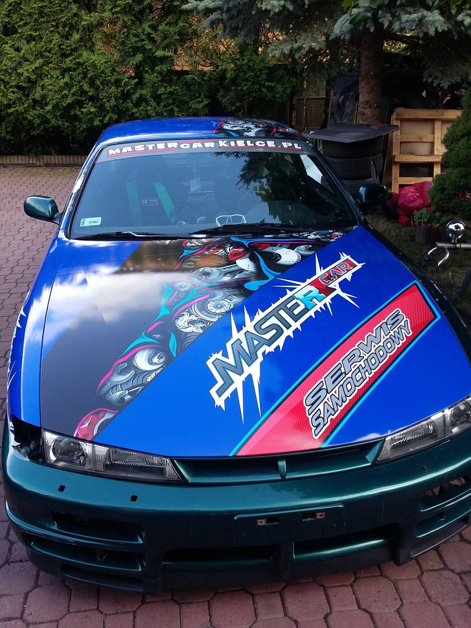 Niebiesko-zielony samochód z grafiką na masce i napisem 'MASTER CAR SERWIS SAMOCHODOWY' na tle zieleni.
