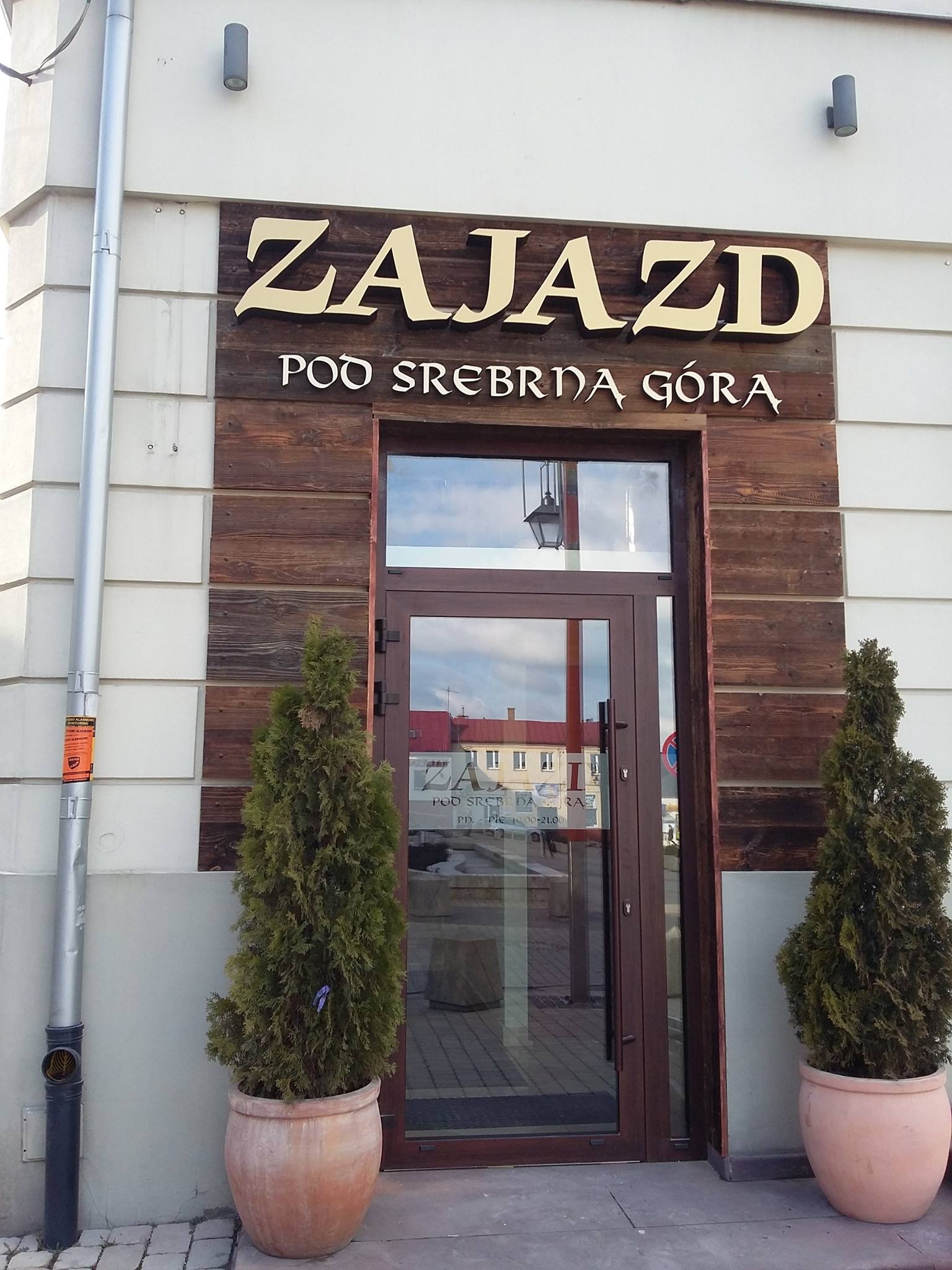 Elewacja budynku z szyldem 'Zajazd Pod Srebrną Górą' nad wejściem, ozdobiona drewnianymi panelami i dwoma tujami w glinianych donicach po bokach drzwi.