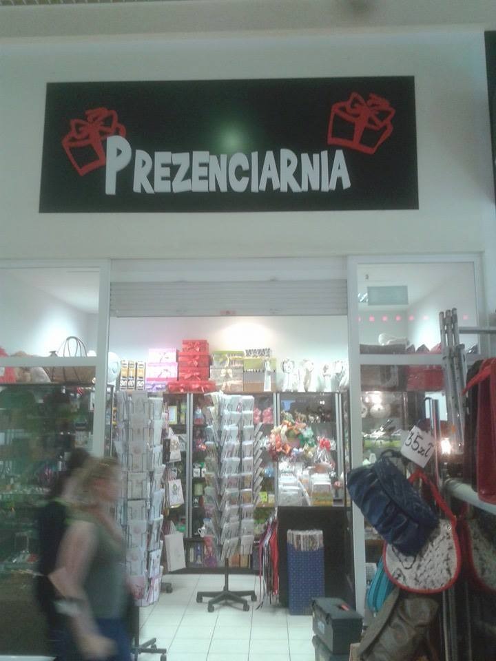 Wnętrze sklepu z prezentami 'Prezenciarnia', widoczny asortyment na półkach i regałach, nad wejściem szyld z nazwą i grafiką prezentów.