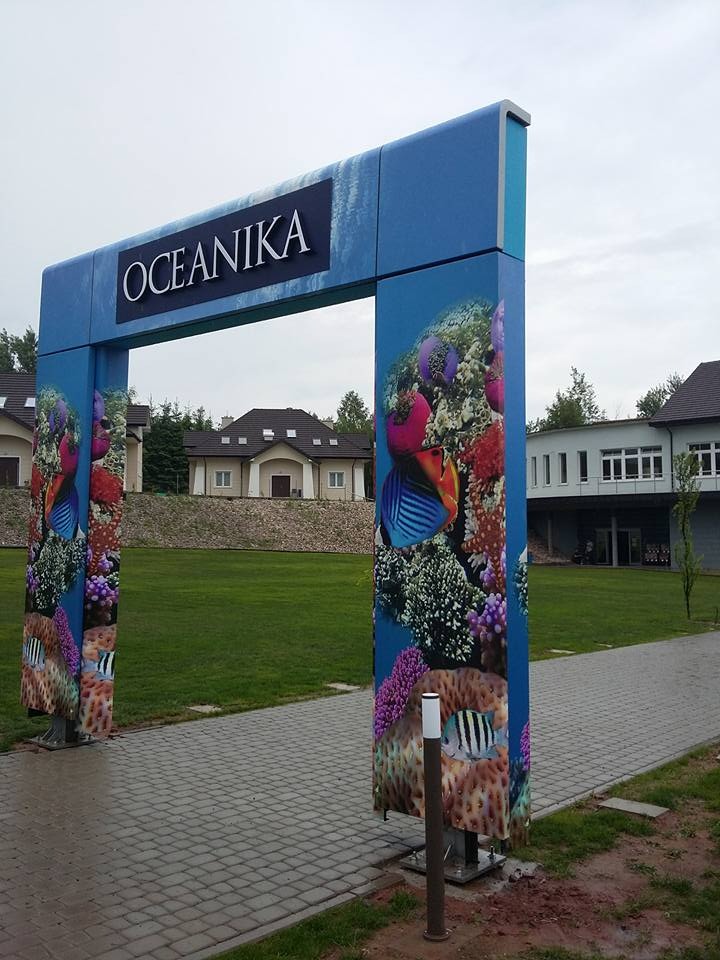 Brama wejściowa z niebieską konstrukcją ozdobioną grafiką rafy koralowej i ryb, nad którą umieszczony jest szyld z napisem 'OCEANIKA', prowadząca do budynku w tle.
