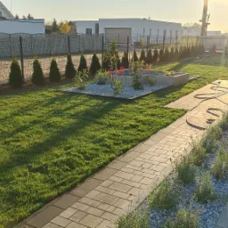 Zadbany ogród przydomowy z równo przystrzyżonym trawnikiem, ozdobnymi tujami wzdłuż ogrodzenia, podwyższoną rabatą z białym żwirem i niskimi roślinami oraz ścieżką z kostki brukowej prowadzącą...