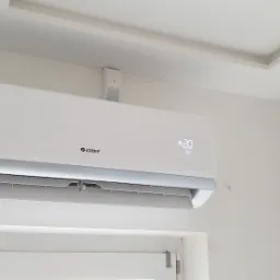 Biała jednostka klimatyzacji marki Gree zamontowana na ścianie nad oknem, wyświetlająca temperaturę 20 stopni Celsjusza.