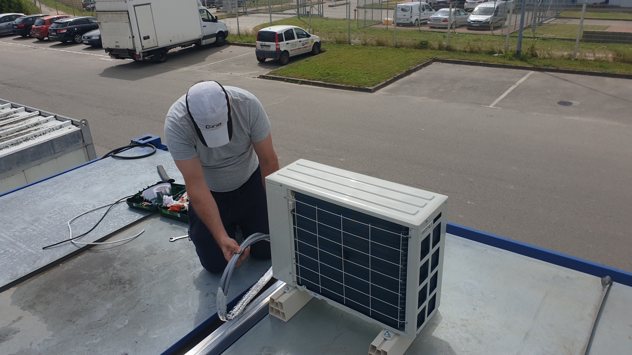 Instalator w trakcie montażu jednostki zewnętrznej klimatyzacji na dachu budynku, widoczna skrzynka z narzędziami i przewód w izolacji.