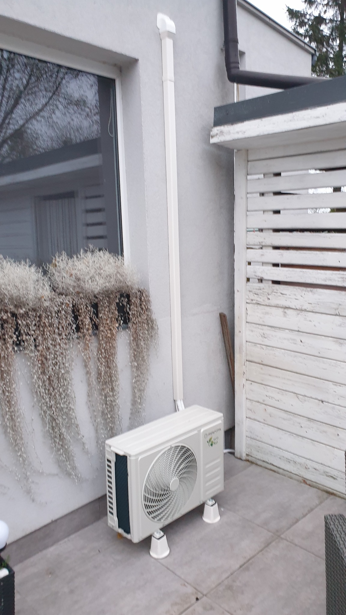Zewnętrzna jednostka klimatyzacji marki Mantra Eco Cool postawiona na białych podkładkach, z widocznymi rurami odprowadzającymi, zamontowana na zewnątrz budynku z białymi ścianami i ozdobnymi...