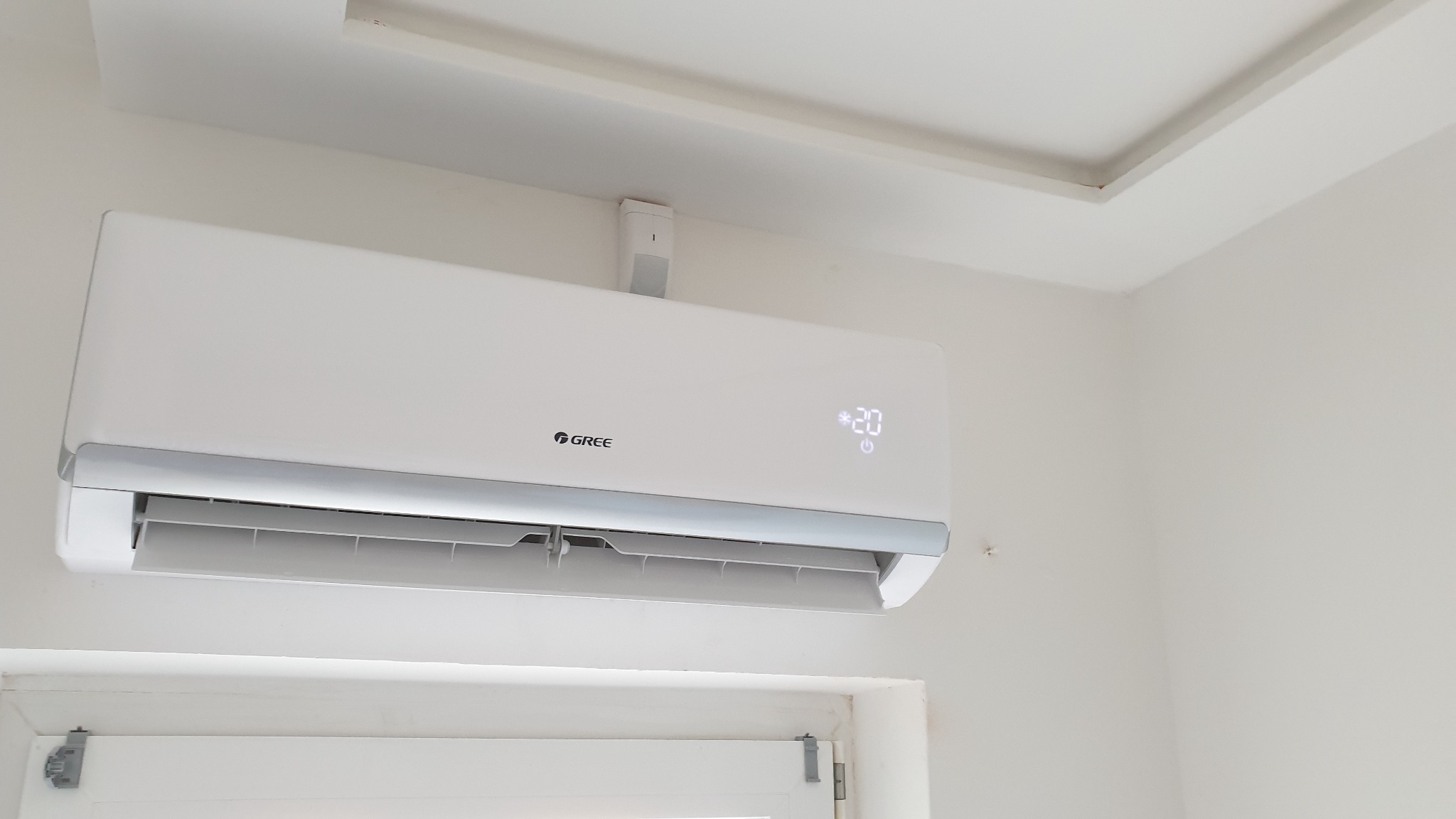 Biała jednostka klimatyzacji marki Gree zamontowana na ścianie nad oknem, wyświetlająca temperaturę 20 stopni Celsjusza.