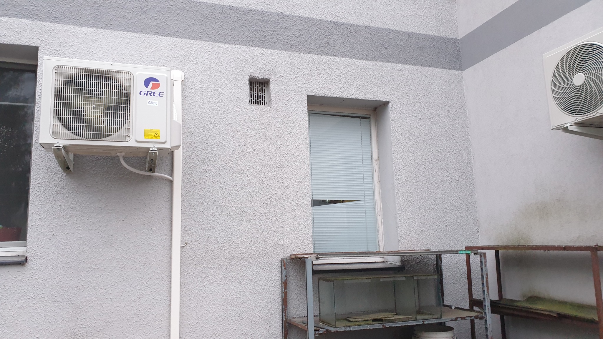 Zewnętrzna jednostka klimatyzacji marki Gree zamontowana na szarej ścianie budynku, widoczna rura odprowadzająca skropliny, obok okno z żaluzjami i metalowy regał z akwarium na dole.