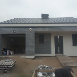 Patbud - Nowoczesny dom jednorodzinny z szarą elewacją i grafitowym dachem. Widoczne materiały budowlane i otwarty garaż. Ujęcie z zewnątrz, w trakcie prac wykończeniowych.