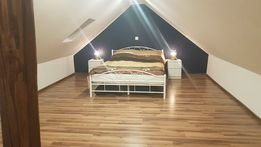 Przytulna sypialnia na poddaszu z drewnianą podłogą, białym łóżkiem i szafkami nocnymi z lampkami. Ściana za łóżkiem pomalowana na ciemny kolor.