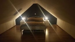Przytulna sypialnia na poddaszu ze skośnym sufitem, oświetlona lampkami nocnymi po bokach łóżka. Ciemne ściany i drewniana podłoga tworzą ciepły klimat.