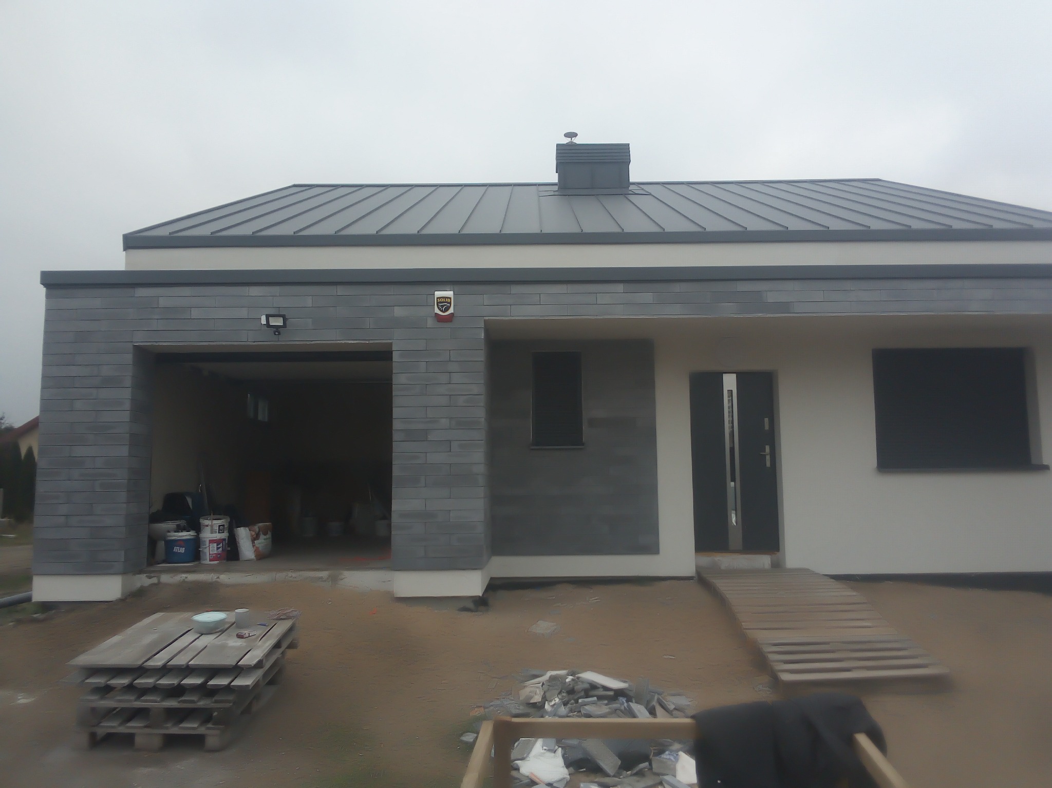 Nowoczesny dom jednorodzinny z szarą elewacją i grafitowym dachem, widoczny garaż z materiałami budowlanymi w środku i paleta przed budynkiem.