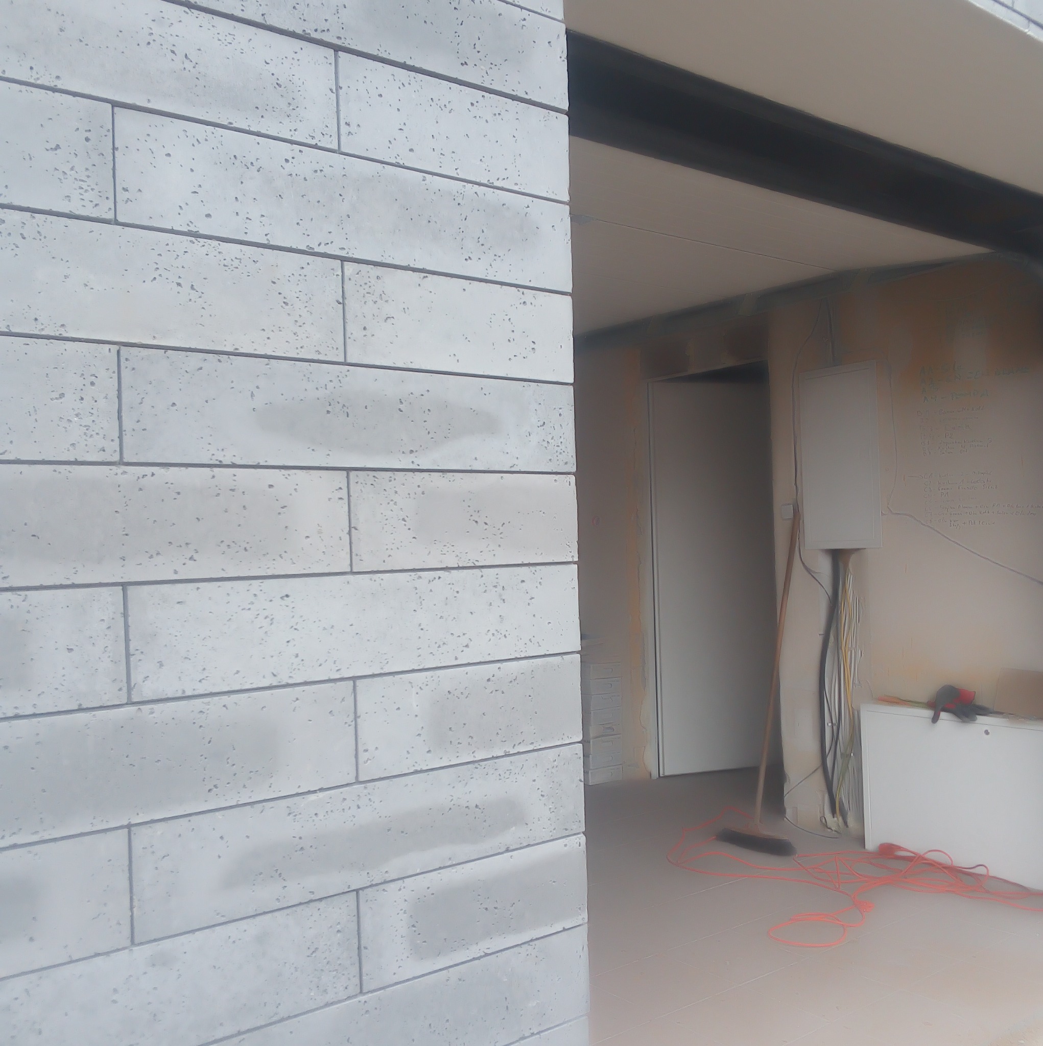 Ściana wykończona szarymi, podłużnymi płytkami imitującymi cegłę. W tle widoczne elementy instalacji elektrycznej i fragment pomieszczenia w trakcie prac wykończeniowych.