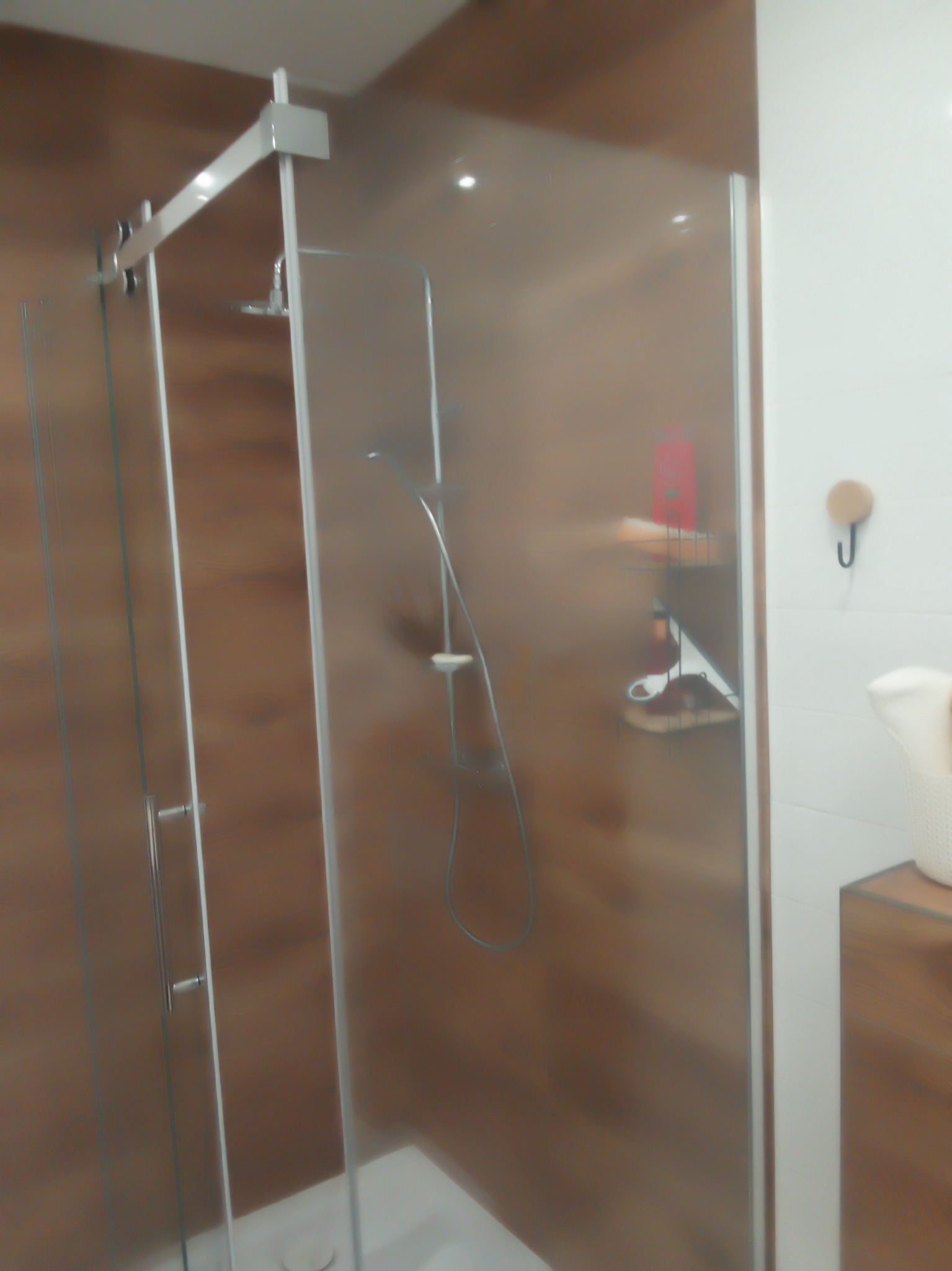 Nowoczesna kabina prysznicowa z przesuwnymi drzwiami, wykończona panelami imitującymi drewno, z półką na kosmetyki i haczykiem na ręcznik.