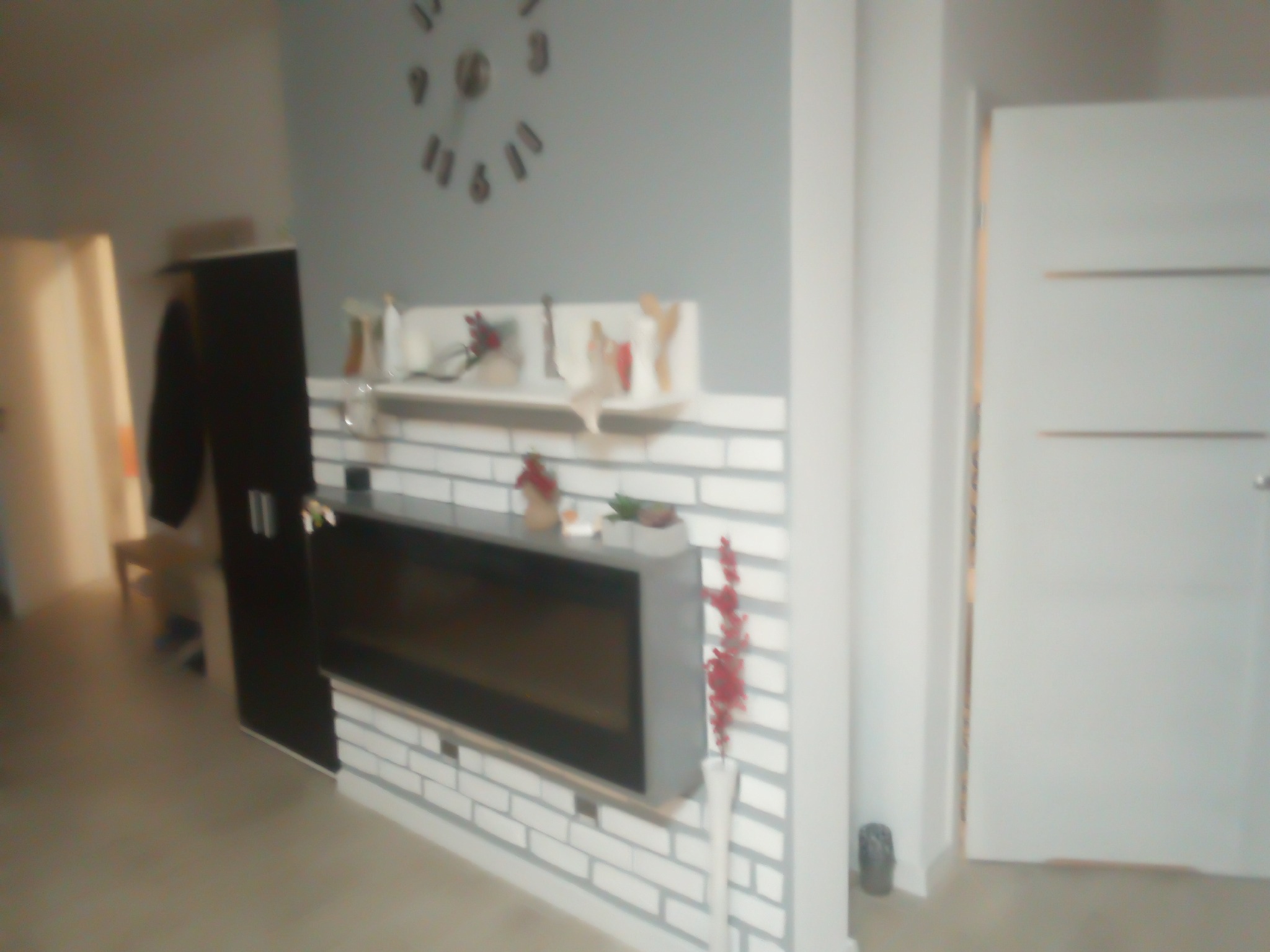 Nowoczesny salon z dekoracyjną ścianą z białej cegły, na której wisi czarny kominek elektryczny. Na ścianie zegar i półka z ozdobami. W tle drzwi i szafa.