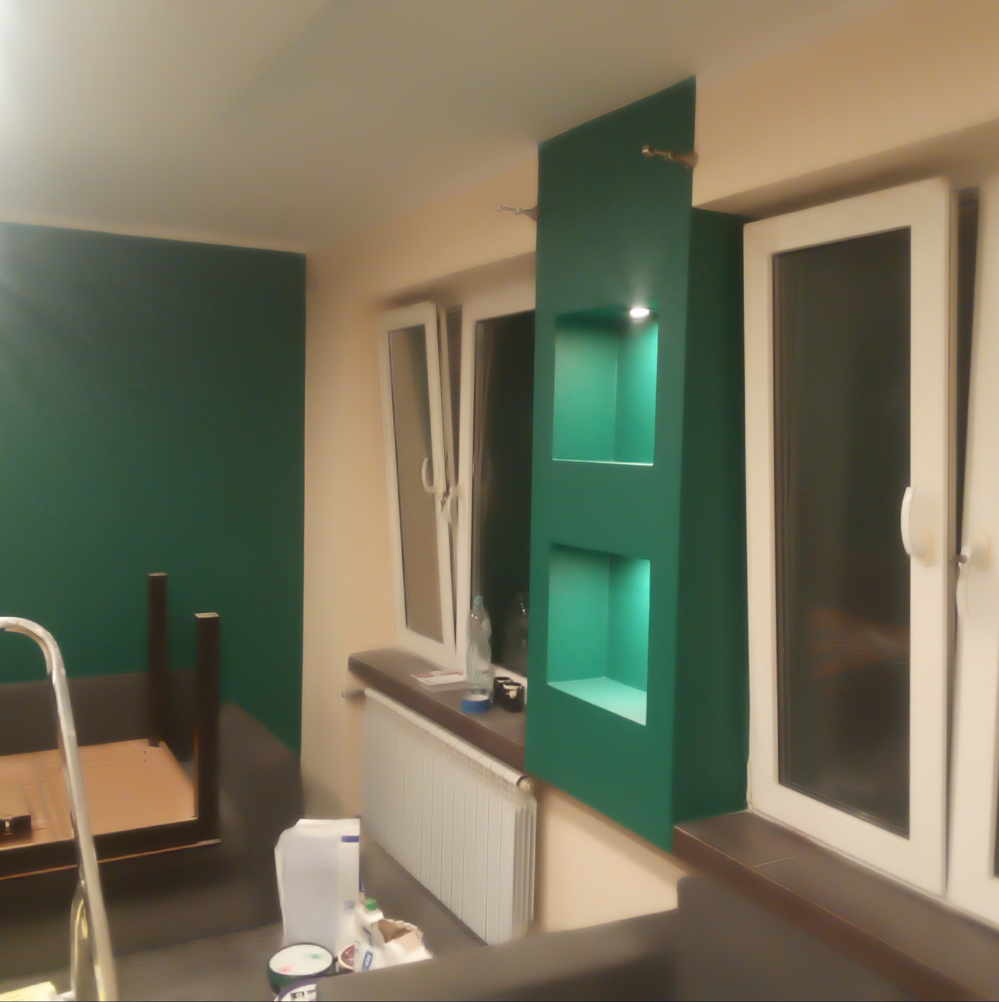 Zielony akcent: ściana i wnęka z oświetleniem LED w odświeżonym wnętrzu. Widoczne okno i drabina malarska, sugerujące prace wykończeniowe.