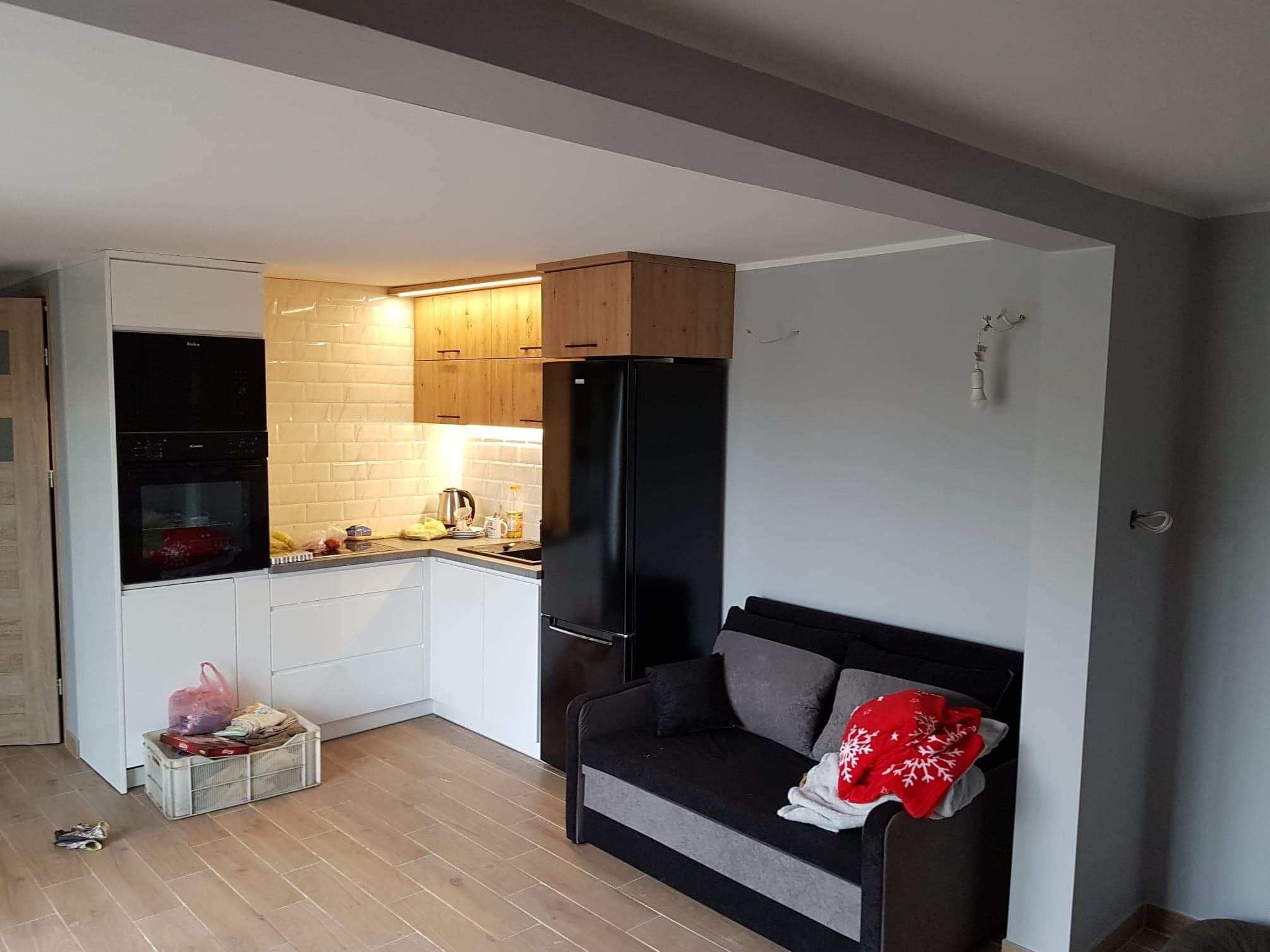 Nowoczesna kuchnia z białymi szafkami, drewnianymi akcentami i czarną lodówką, z widoczną częścią salonu z szarą sofą i drewnianą podłogą.