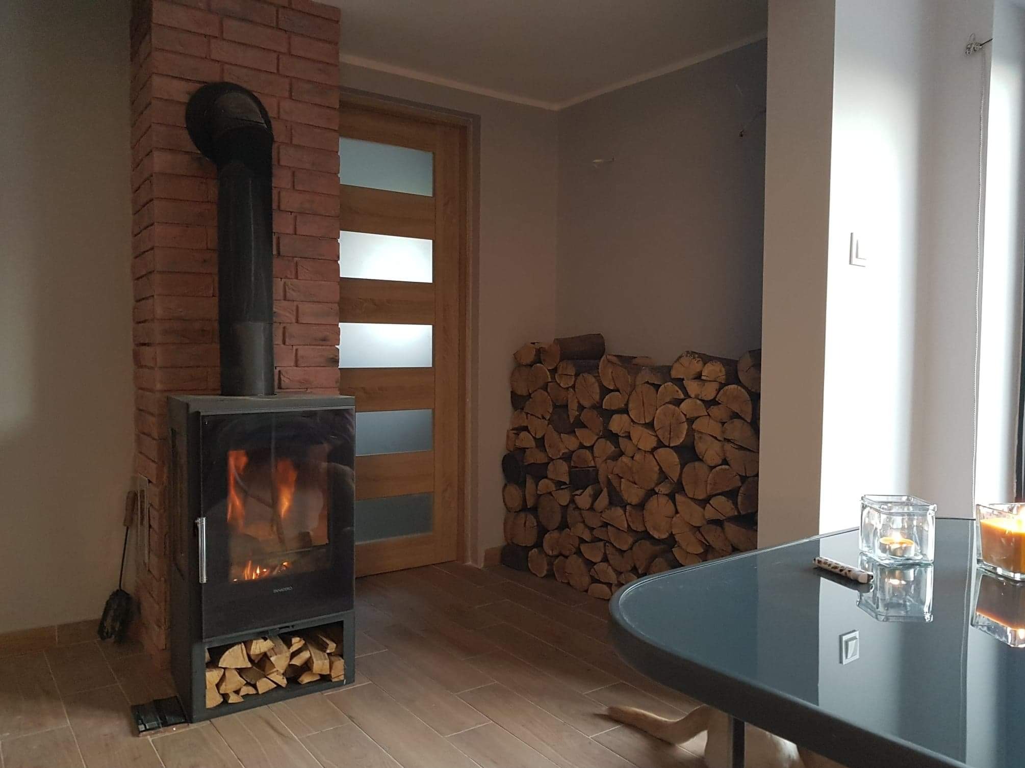 Nowoczesny salon z kominkiem obudowanym czerwoną cegłą, drewnem ułożonym przy ścianie i szklanym stolikiem kawowym. Parkiet w odcieniach brązu.