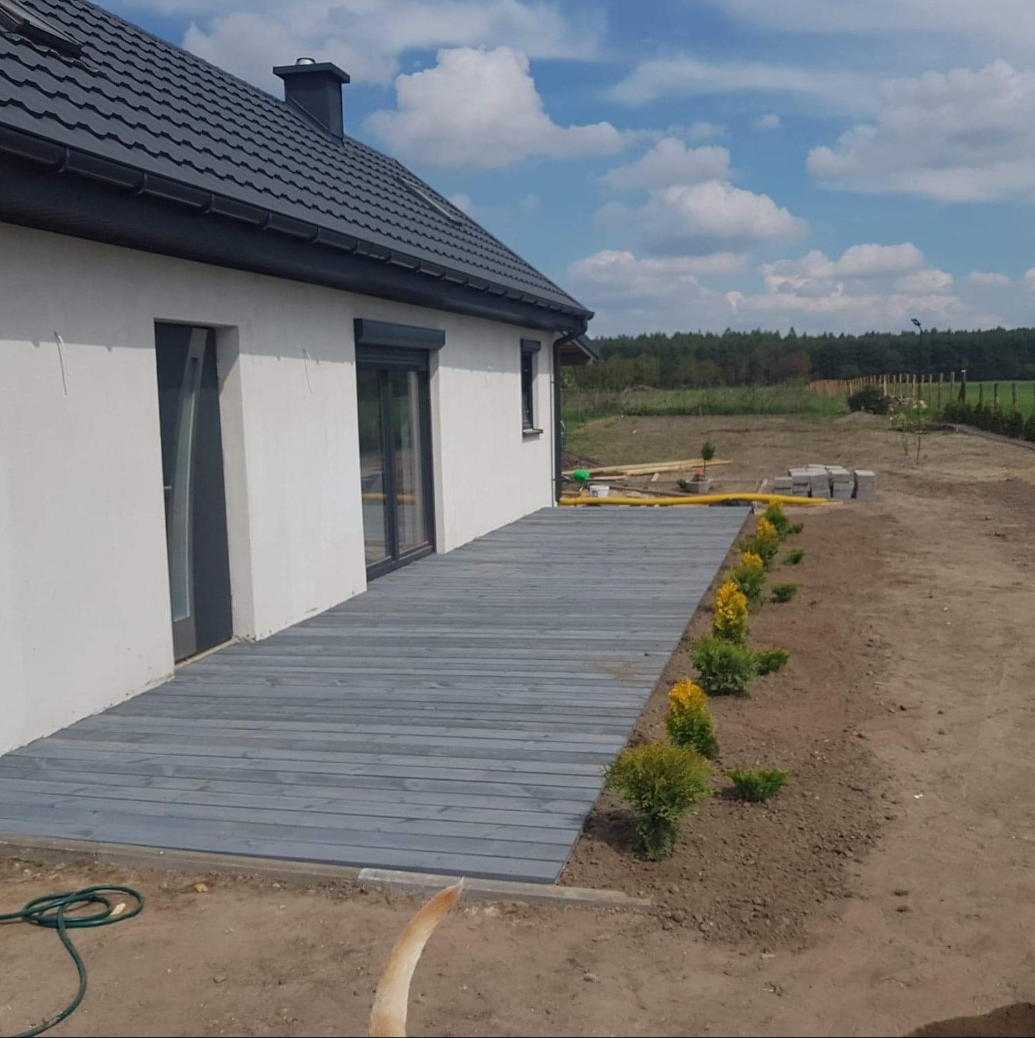 Szara drewniana podłoga tarasu przy białym domu z szarą stolarką, świeżo posadzone tuje, w tle las i błękitne niebo z chmurami.