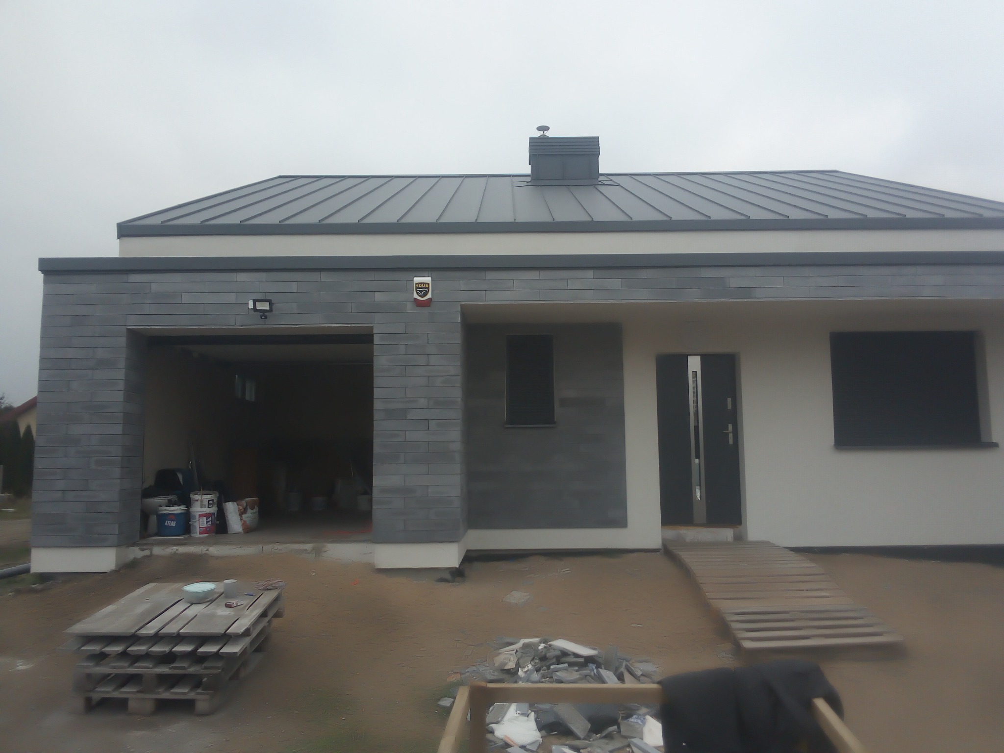 Nowoczesny dom jednorodzinny z szarą elewacją i grafitowym dachem. Widoczne materiały budowlane i otwarty garaż. Ujęcie z zewnątrz, w trakcie prac wykończeniowych.