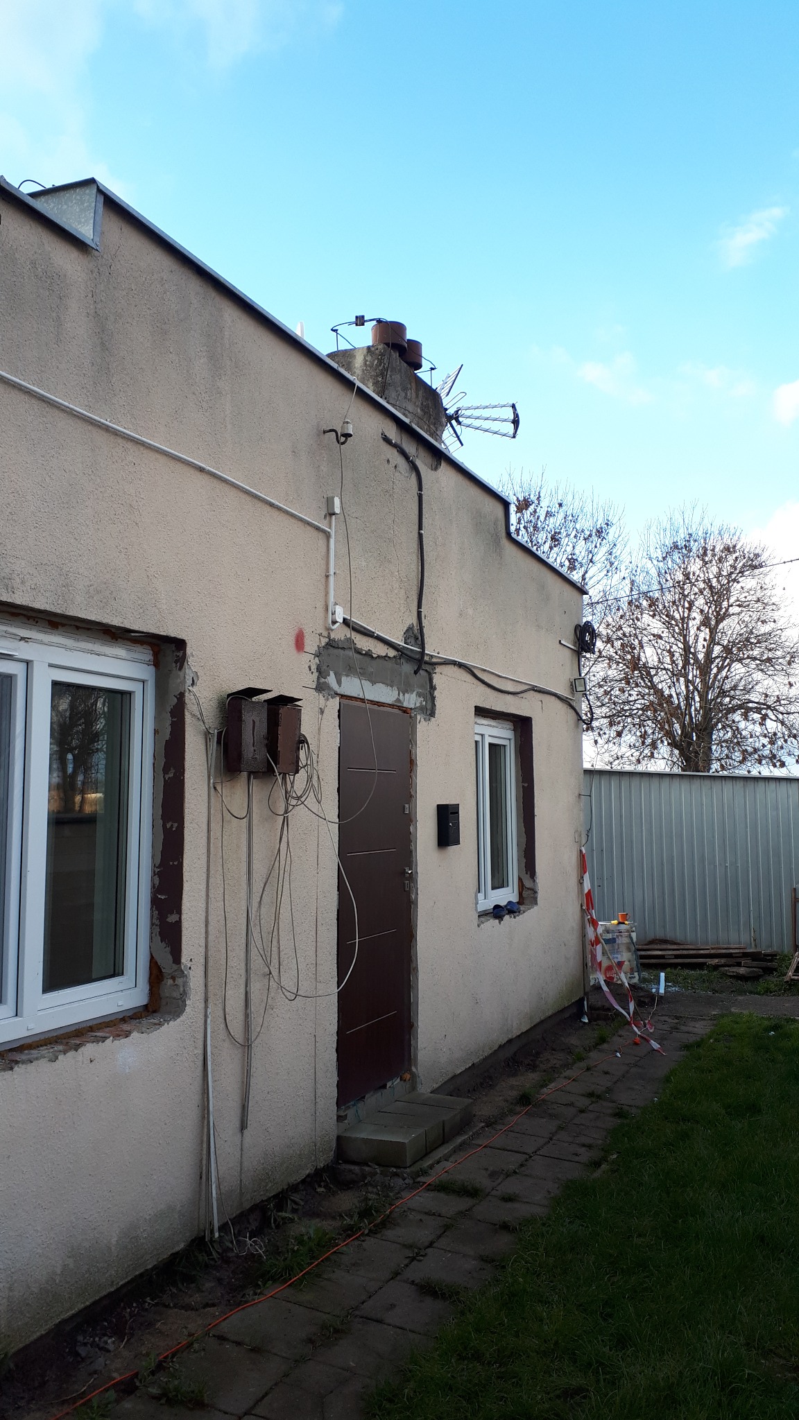 Elewacja domu w trakcie prac: nowe okna, świeże obramowania, widoczne przewody i skrzynki elektryczne. Fragment chodnika i trawnika. Błękitne niebo w tle.