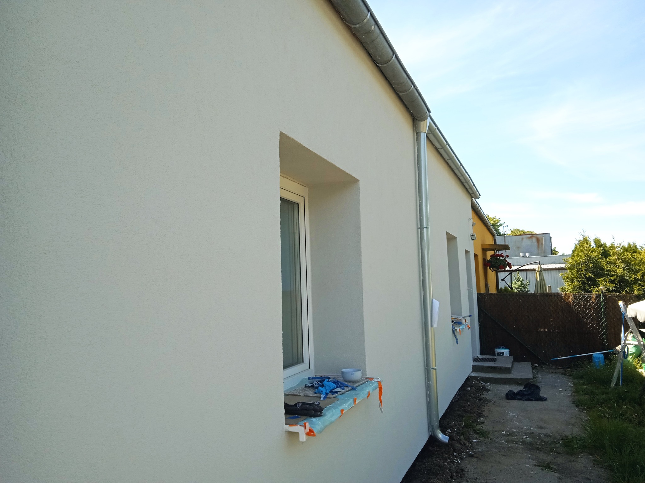 Otynkowana elewacja budynku jednorodzinnego w jasnym odcieniu, z widocznymi oknami i rynnami. Na parapecie okna leżą narzędzia i akcesoria malarskie.