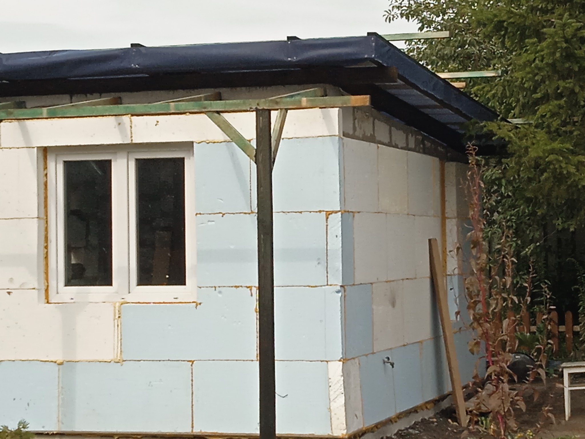 Budynek w trakcie ocieplania styropianem, widoczne okno i drewniana konstrukcja dachu. Etap prac przed wykończeniem elewacji.