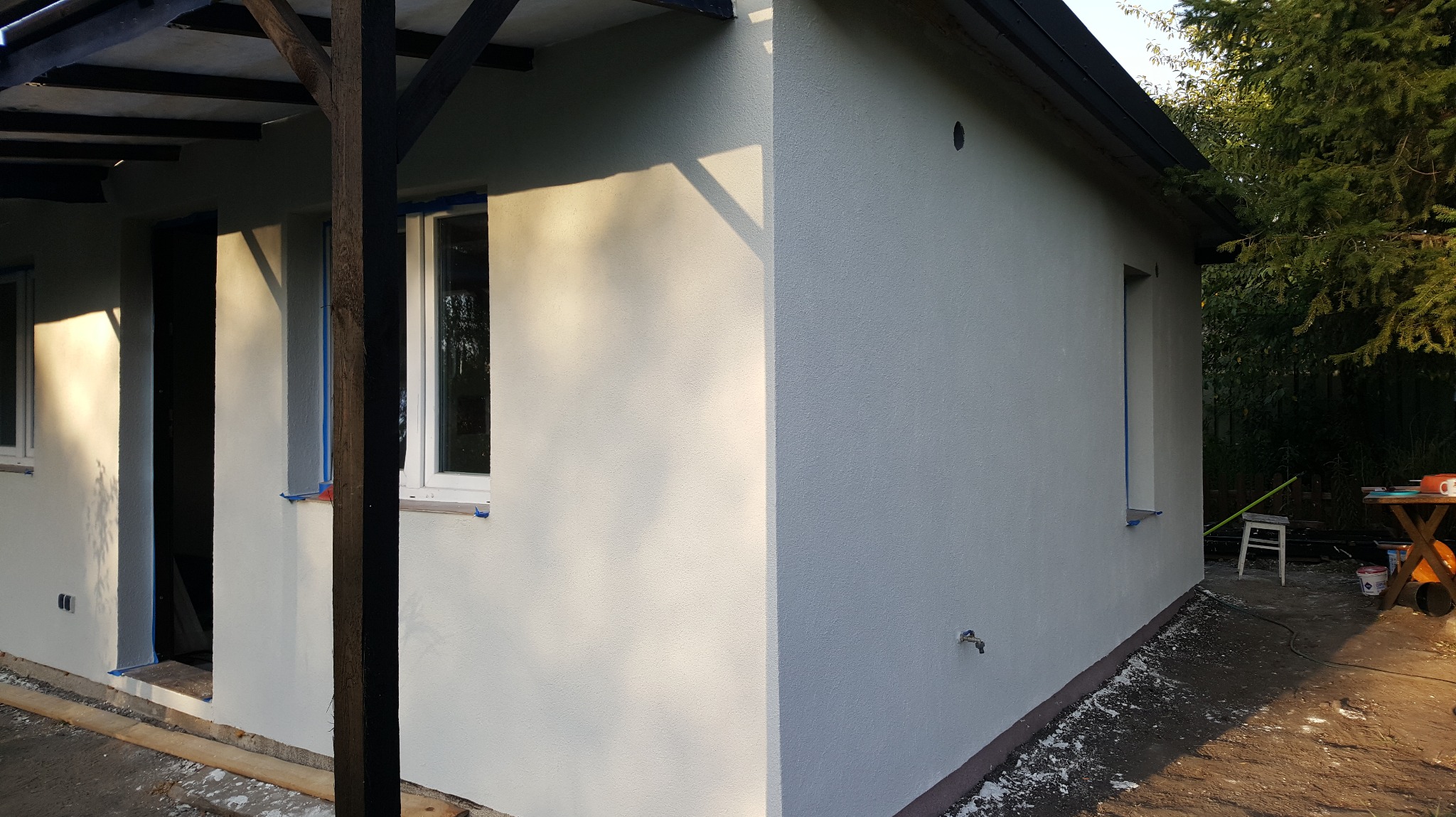 Otynkowana elewacja budynku z oknami zabezpieczonymi taśmą malarską. Widoczne ślady prac wykończeniowych na ziemi. Ujęcie z zewnątrz, w słoneczny dzień.