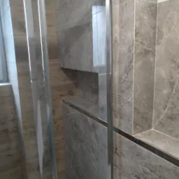 Szklana kabina prysznicowa z przesuwnymi drzwiami, wykończona szarymi płytkami imitującymi marmur z półką wnękową.