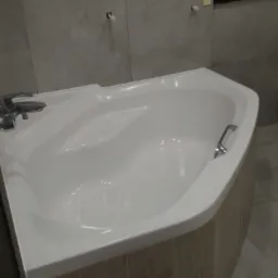 Biała wanna narożna z obudową imitującą drewno, chromowana bateria i uchwyt, szare płytki na ścianach i podłodze, widoczne niedokończone punkty instalacji.
