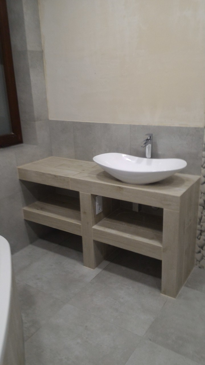 Nowoczesna łazienka z umywalką nablatową o owalnym kształcie, zamontowaną na szafce z jasnego drewna z półkami, widoczna instalacja elektryczna, szara płytka na podłodze i ścianach.