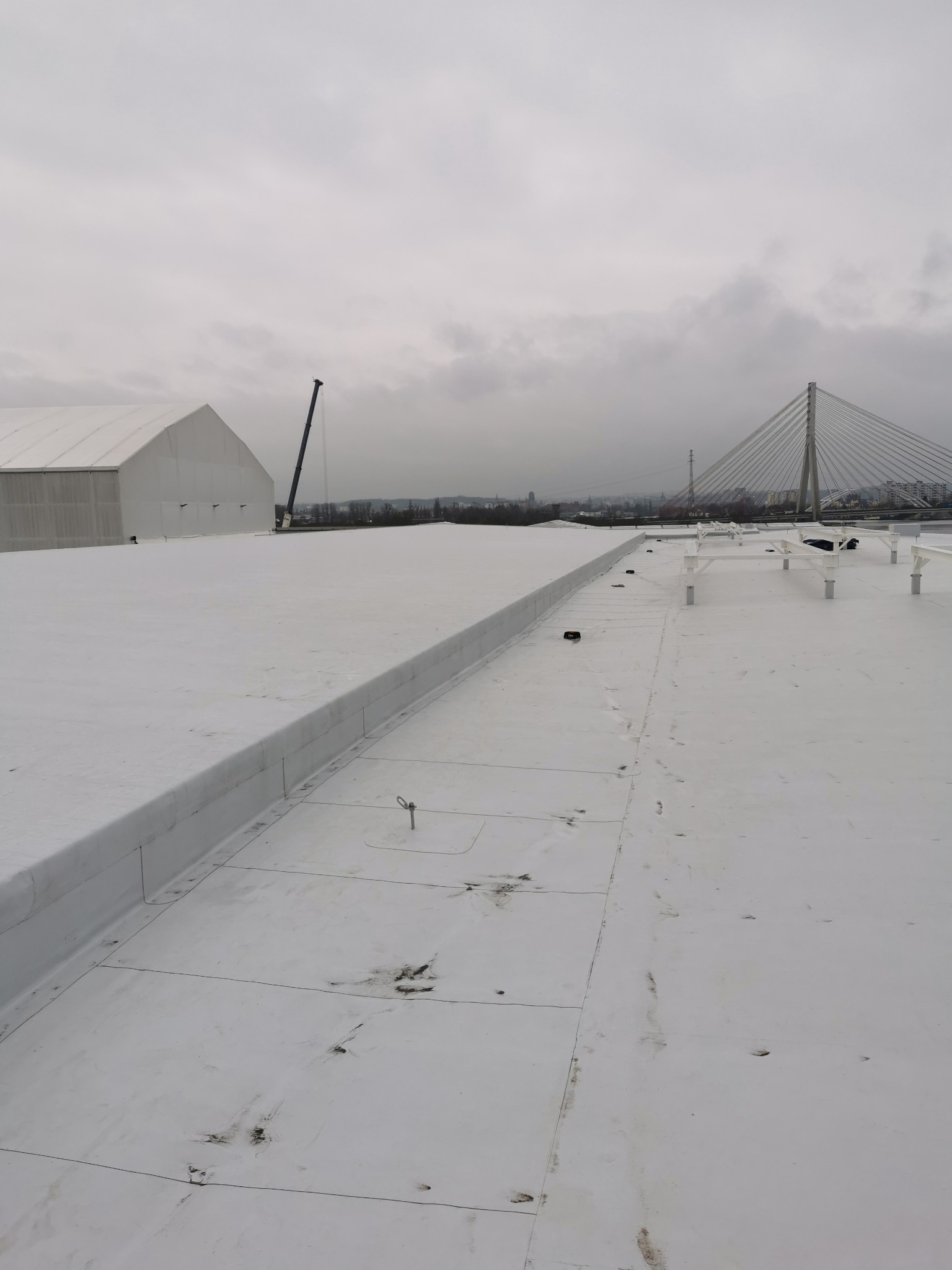 Biały dach płaski z widocznymi panelami i elementami mocującymi, na tle pochmurnego nieba, budynku magazynowego i mostu wiszącego w oddali.