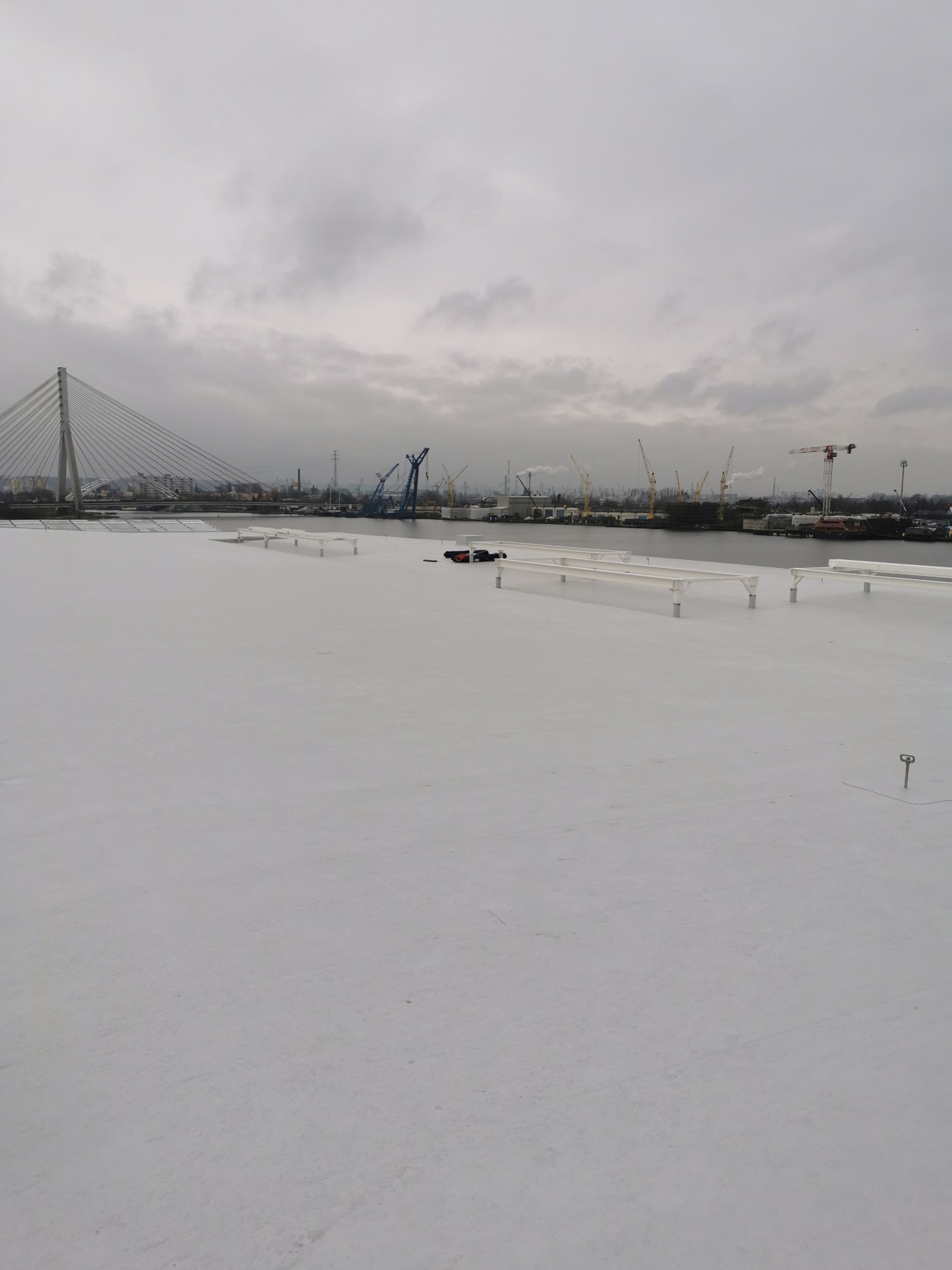 Biały, płaski dach pokryty membraną, widoczne elementy mocujące, w tle panorama portu z dźwigami i mostem wantowym w pochmurny dzień.