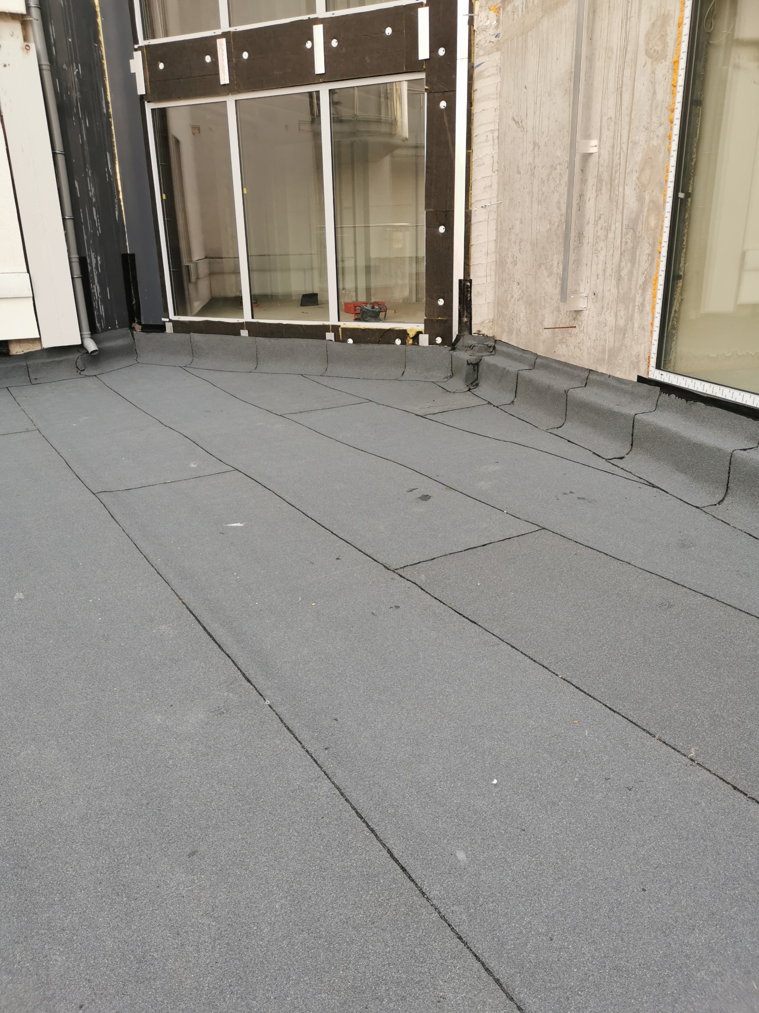 Widok z góry na płaski dach pokryty papą termozgrzewalną, z widocznymi obróbkami blacharskimi przy oknach i ścianie budynku.