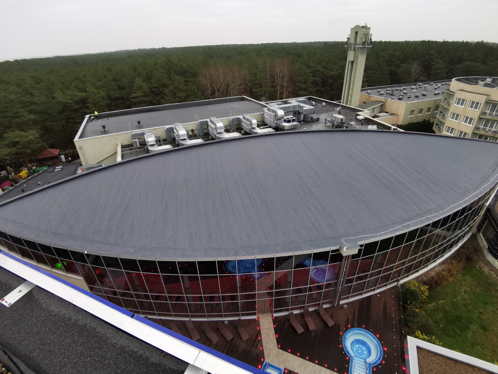 Widok z góry na duży, płaski, szary dach budynku z zaokrągloną fasadą, widoczne systemy wentylacyjne na dachu, las w tle i fragment konstrukcji innego dachu na pierwszym planie.