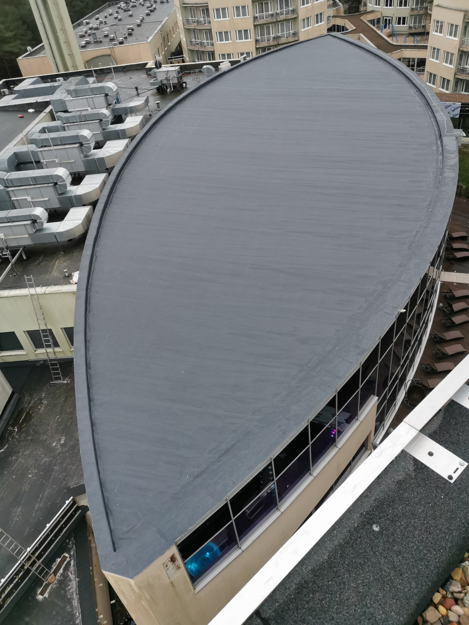 Widok z góry na płaski, szary dach budynku o nietypowym, zaokrąglonym kształcie, przylegający do budynku z oknami i tarasem z leżakami, w tle inne budynki i instalacje wentylacyjne.