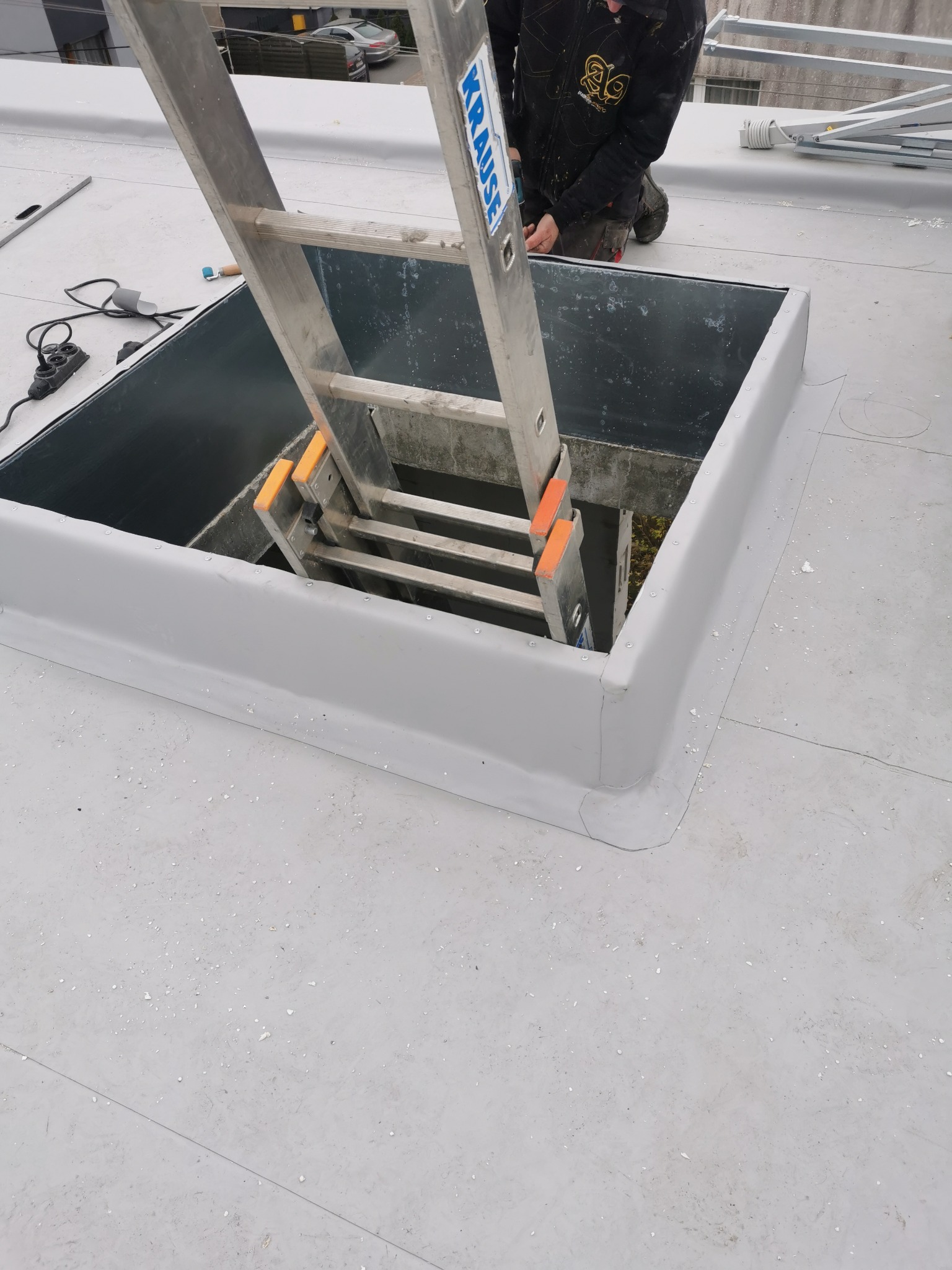 Szary, płaski dach z wyłazem, zabezpieczony obróbką blacharską, z przystawioną drabiną Krause. Widoczny fragment pracownika w kombinezonie.