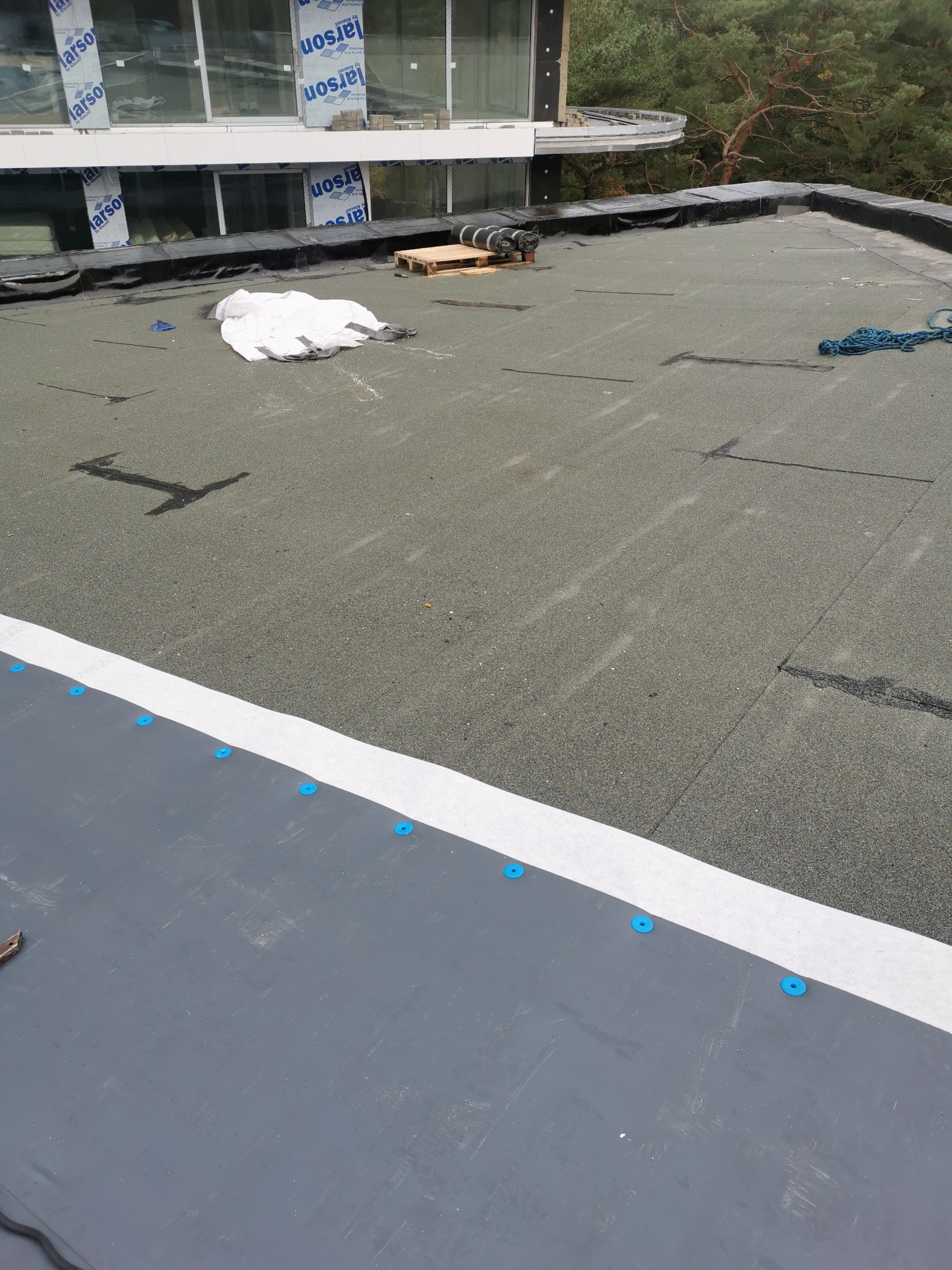 Nowoczesny dach płaski z papy termozgrzewalnej w trakcie prac wykończeniowych, widoczne rolki papy, paleta, worki z materiałem, niebieskie mocowania i detale obróbek blacharskich.