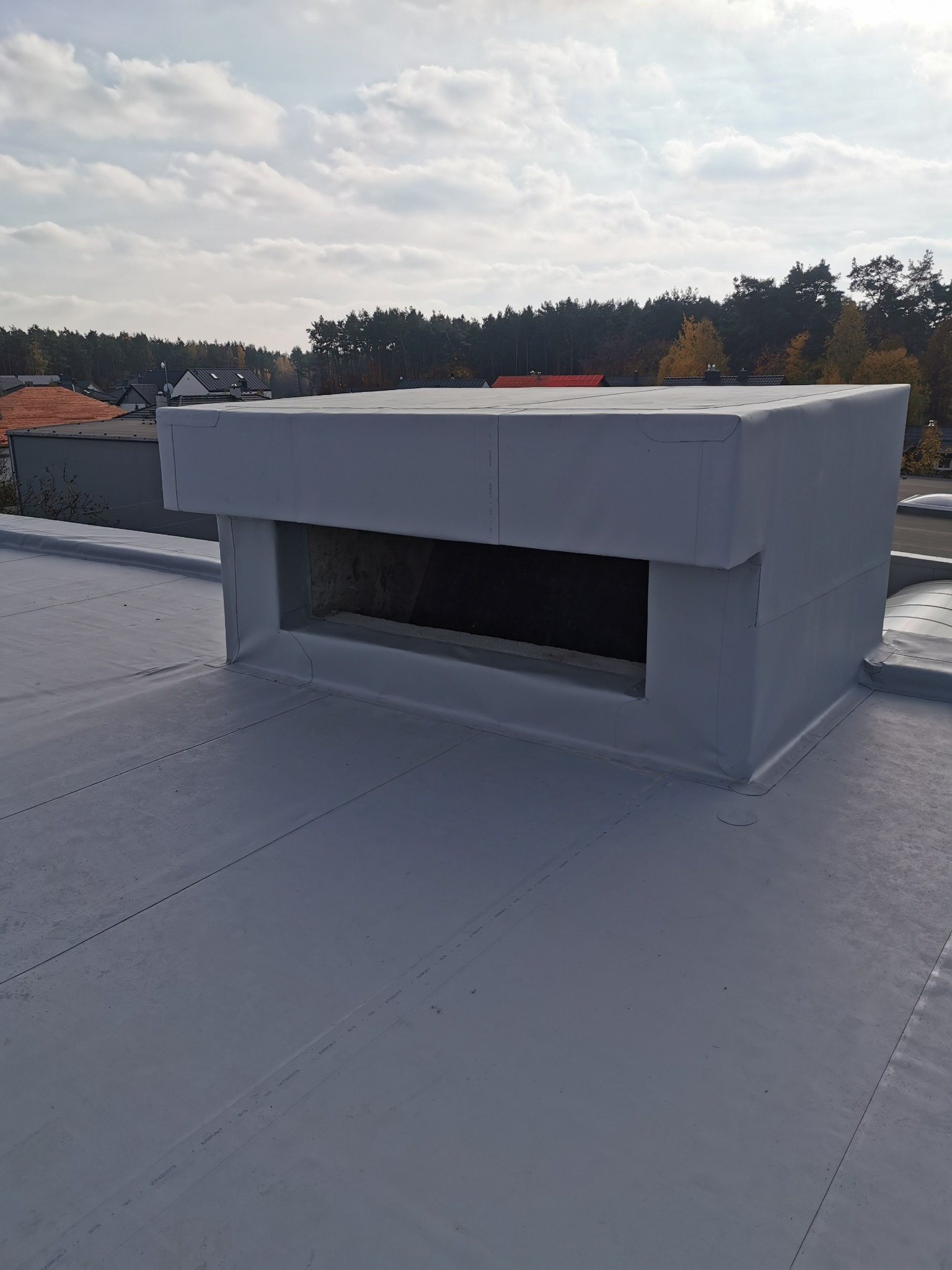 Szary dach pokryty membraną z wykończonym kominem wentylacyjnym w kształcie prostopadłościanu; widok z góry na tle drzew i pochmurnego nieba.