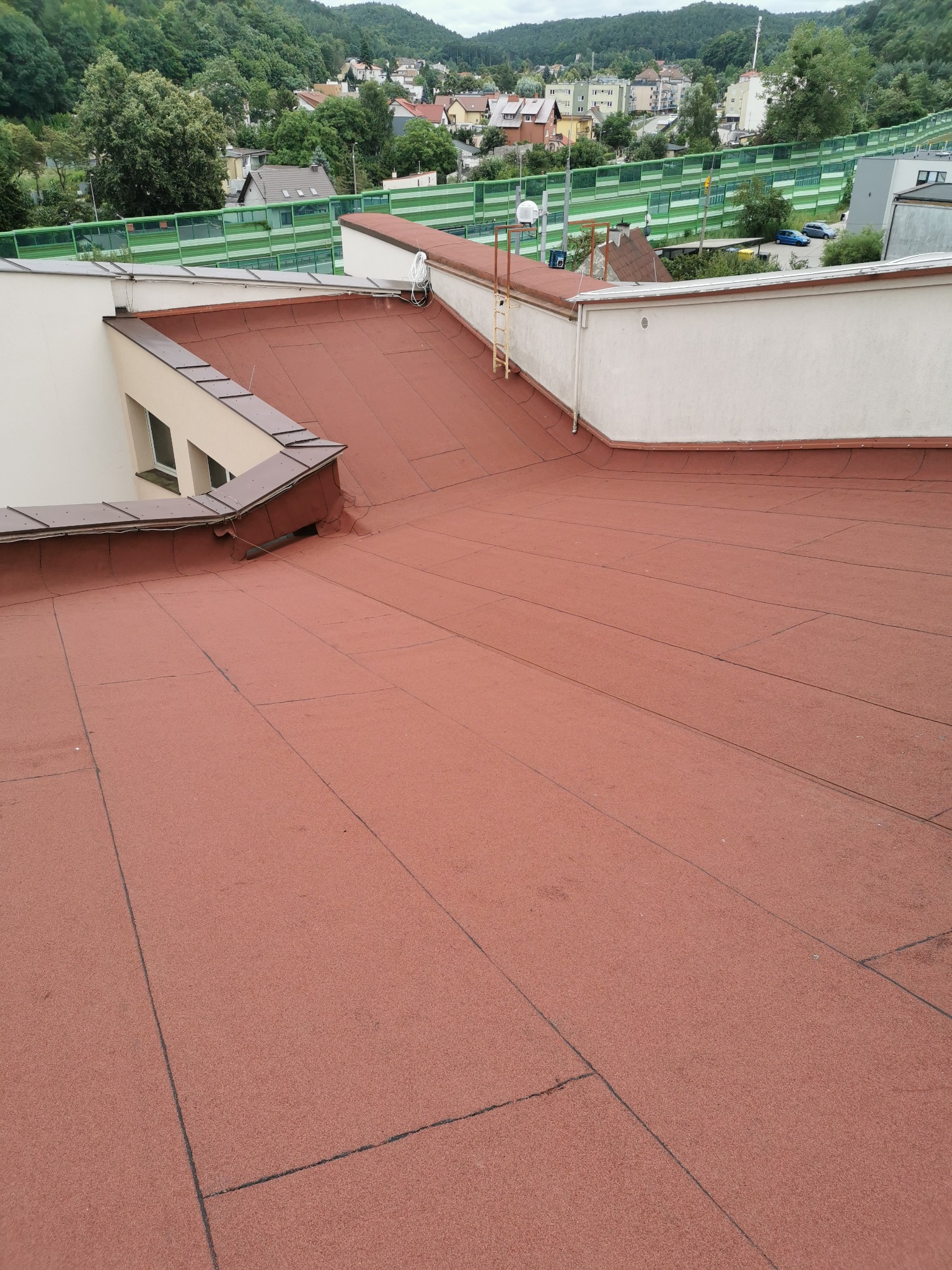 Płaski dach pokryty czerwoną papą termozgrzewalną, widoczne łączenia papy, fragment ściany budynku z oknami oraz drabina przy krawędzi dachu, w tle zabudowa miejska i zalesione wzgórza.