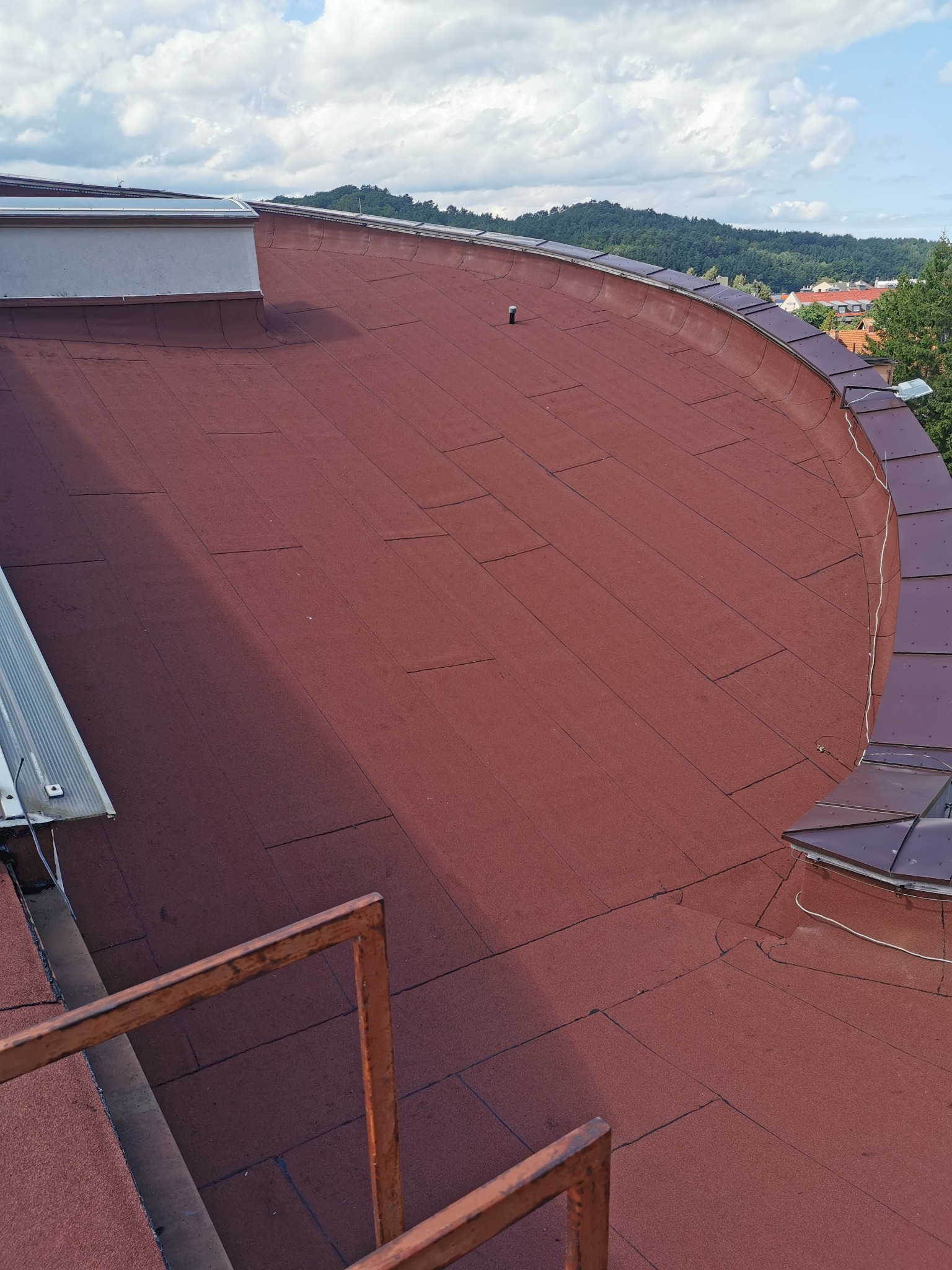 Widok z góry na płaski dach pokryty papą w kolorze ceglastym, z widocznymi obróbkami blacharskimi na krawędziach i elementami infrastruktury dachowej, na tle zabudowań i zalesionych wzgórz...