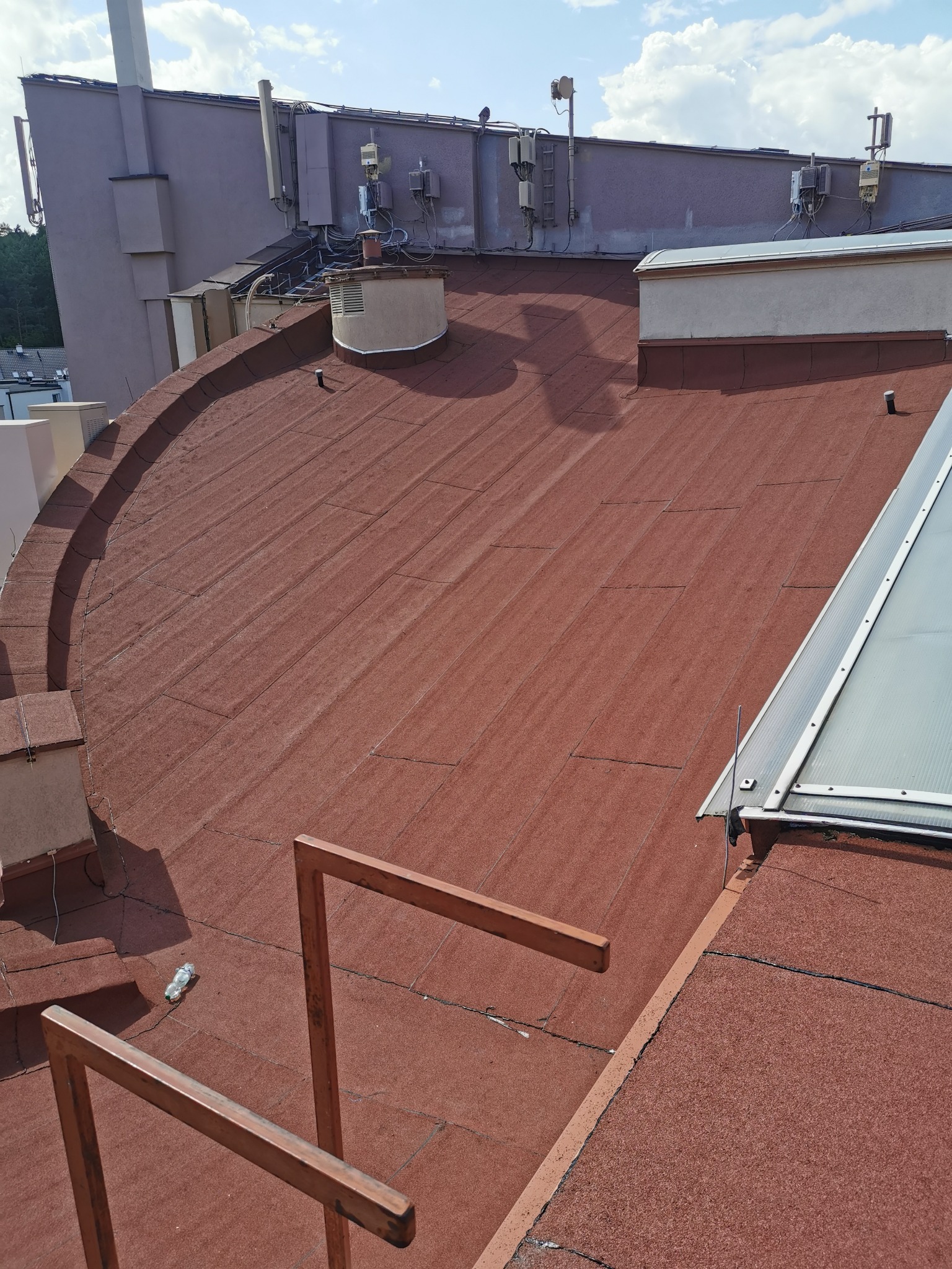 Czerwony dach pokryty papą termozgrzewalną z widocznym świetlikiem dachowym i elementami instalacji na budynku w tle; na dachu leży pusta butelka PET