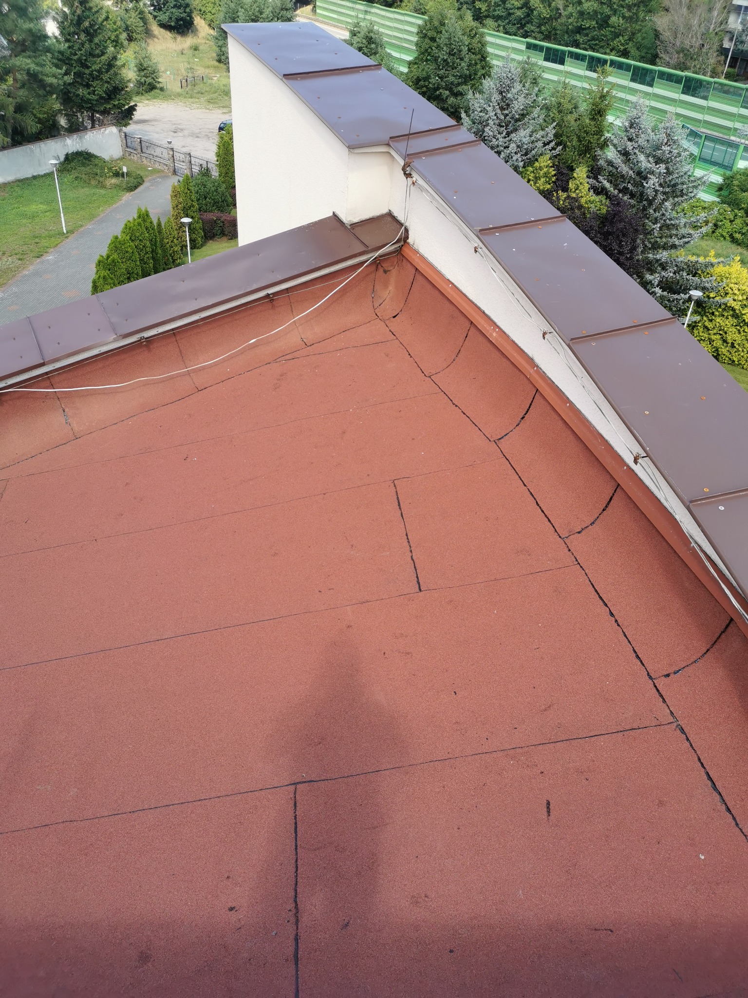 Widok z góry na fragment dachu pokrytego czerwoną papą bitumiczną, z widocznymi brązowymi obróbkami blacharskimi przy krawędzi i białym przewodem biegnącym wzdłuż nich. W tle zieleń drzew...