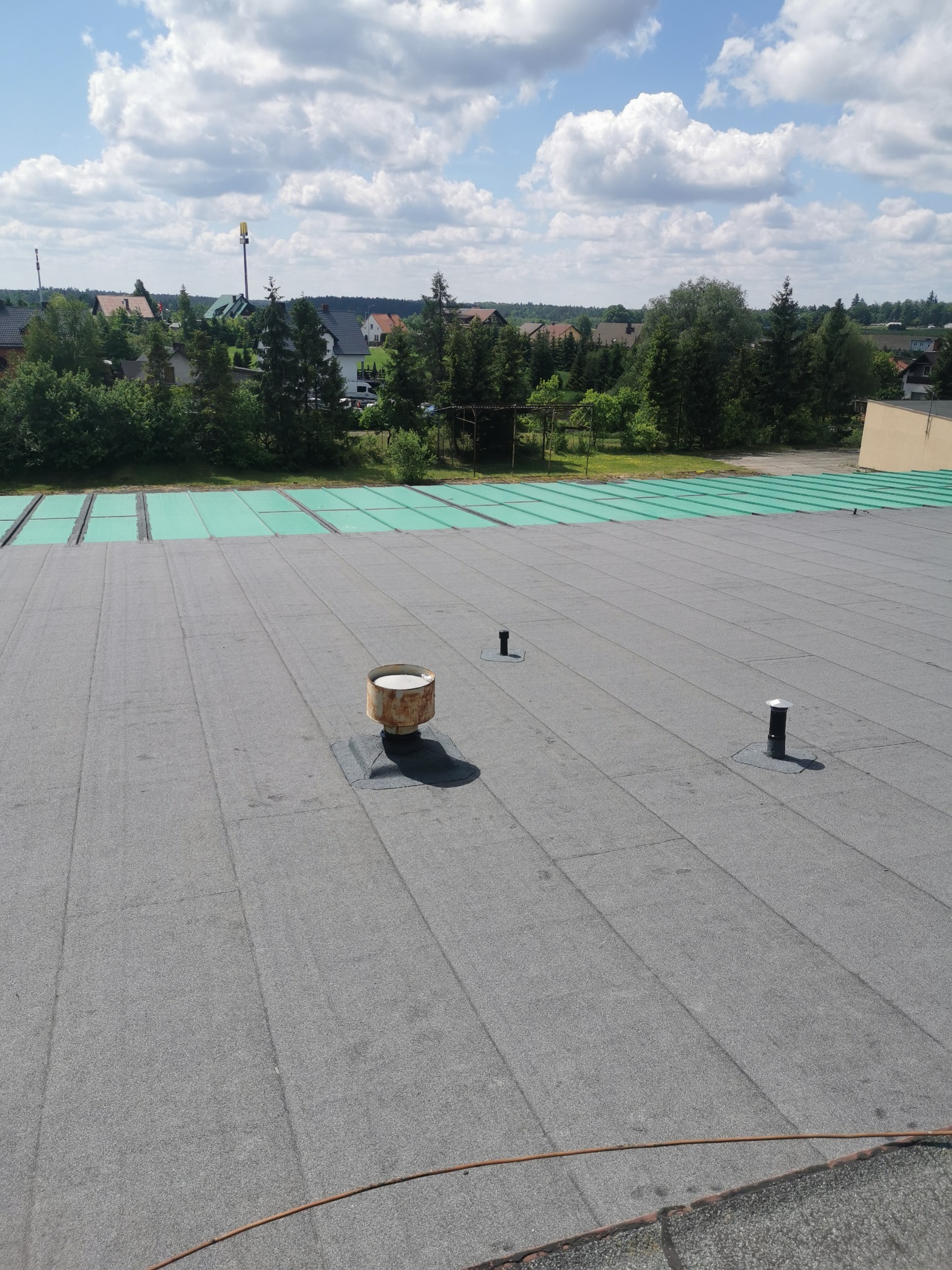 Widok z góry na płaski dach pokryty papą, z widocznymi kominkami wentylacyjnymi i fragmentami zielonej izolacji termicznej, w tle zabudowa jednorodzinna i błękitne niebo z obłokami.