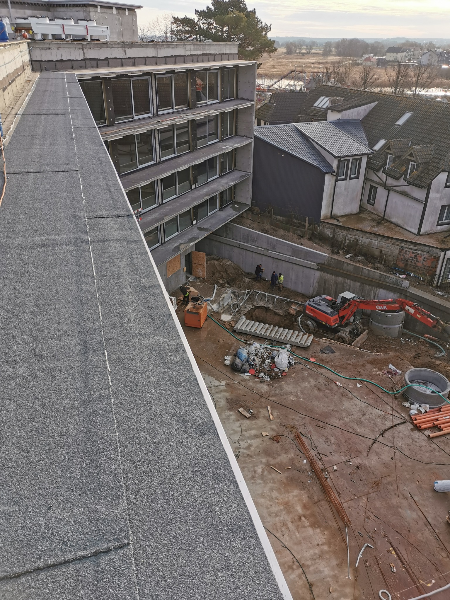 Widok z góry na płaski dach pokryty papą, z widocznym budynkiem w trakcie budowy, wykopem i pracującym sprzętem budowlanym, a w tle zabudowa jednorodzinna.