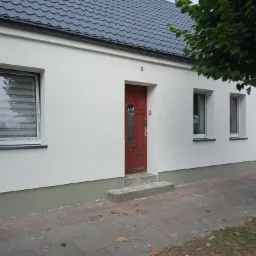 Odnowiona elewacja budynku jednorodzinnego z nowym tynkiem w kolorze białym, z brązowymi oknami i drzwiami wejściowymi, oraz szarym cokołem i dachem z dachówki ceramicznej.