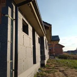 Ściana budynku w trakcie ocieplania styropianem grafitowym, z widocznymi fragmentami pianki montażowej i zamontowaną rynną spustową. W tle widać kolejny budynek w stanie surowym otwartym.