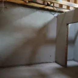 Wnętrze pomieszczenia w surowym stanie po nałożeniu tynków, widoczne niedoskonałości powierzchni i otwór drzwiowy bez ościeżnicy, drewniana konstrukcja stropu z instalacjami pod sufitem.